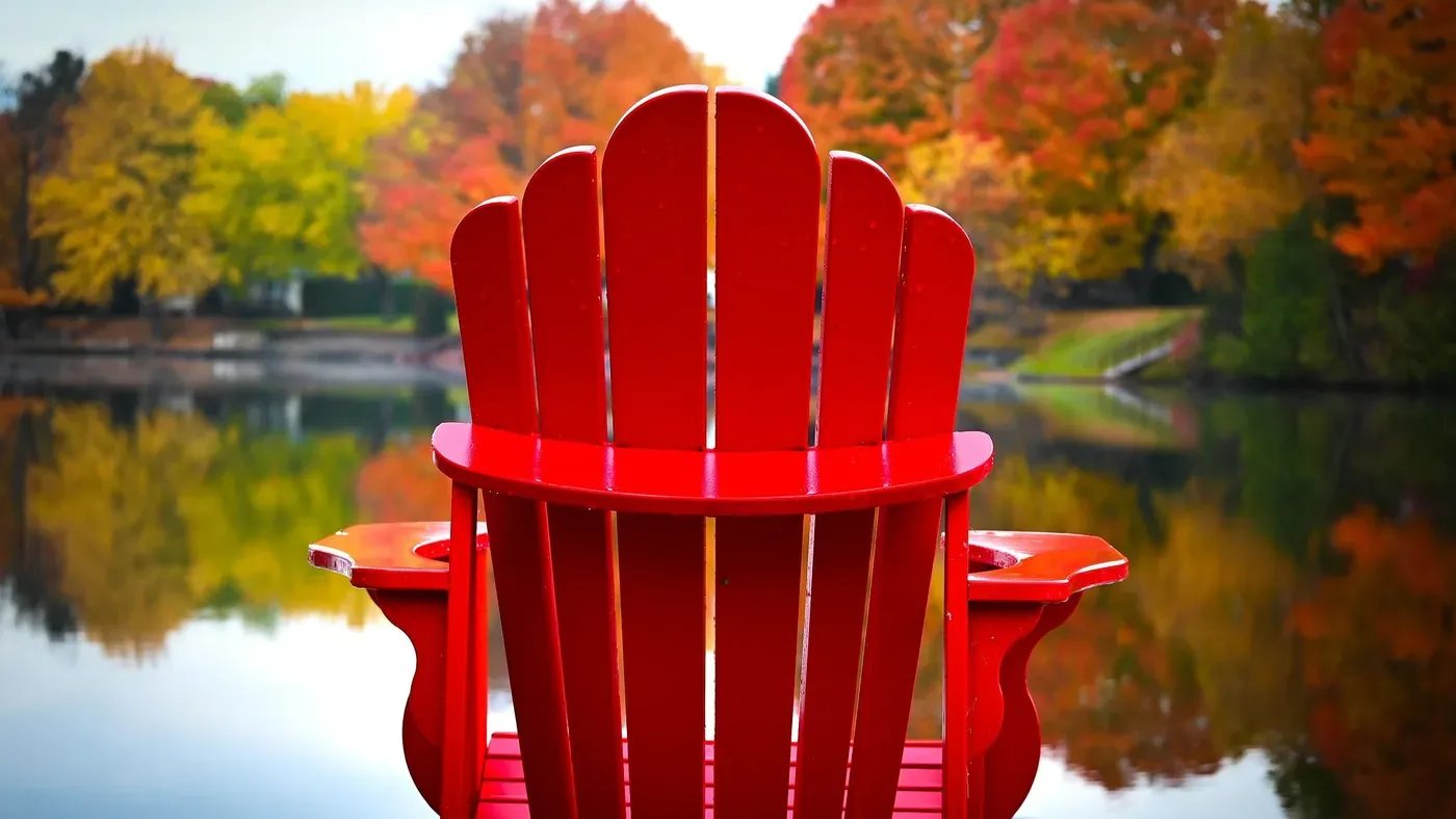 Red Muskoka chair on a dock facing a lake lined with autumn colour, the season for closing up a water-access cottage