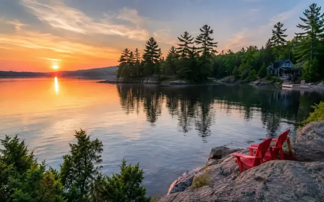 Two red Muskoka chairs on a granite point at sunset, with a cottage among the pines across the water