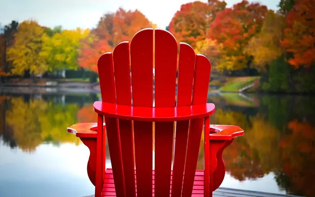 Red Muskoka chair on a dock facing a lake lined with autumn colour, the season for closing up a water-access cottage